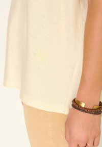 Gros plan sur la main d'une personne portant un bracelet en cuir tressé marron avec des perles en bois et une plaque en or, sur des vêtements beiges.