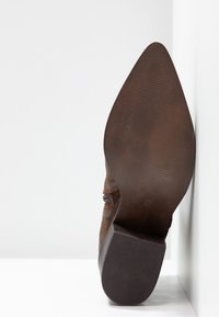 Hnědé nubukové kotníkové boty s špičatou špičkou a vrstvovým podpatkem. Podešev má drsný povrch a plochý, zaoblený okraj. Velikost označena jako 37.