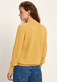 Frau mit welligem braunem Haar, die einen senfgelben strukturierten Langarmpullover und blaue Jeans trägt, von links auf einem einfarbigen Hintergrund posiert.