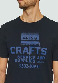 Zwart T-shirt met korte mouwen met gedurfde blauwe grafische tekst "ARTS & CRAFTS SERVICE AND SUPPLIES," met een ontwerp van een verfroller.