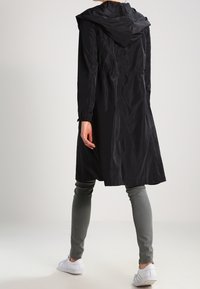 Černý kabát s kapucí vyrobený z lehkého, lesklého materiálu s nastavitelným pasem, dosahující do poloviny lýtek, doplněný šedými kalhotami a bílými teniskami.