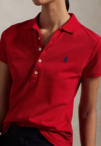 Kvinde iført en rød kortærmet polo-shirt med hvide knapper og en lille blå broderet logo på venstre bryst.