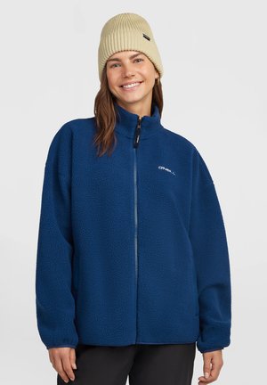 Glimlachende vrouw met een beige gebreide muts, een blauwe full-zip fleecejas en een zwarte broek tegen een effen lichtgrijze achtergrond.