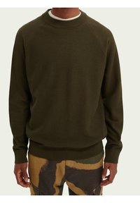 Man draagt een olijfgroene trui met ronde hals en een pantalon met een patroon in groen, beige en zwart camouflageontwerp, staand tegen een effen achtergrond.