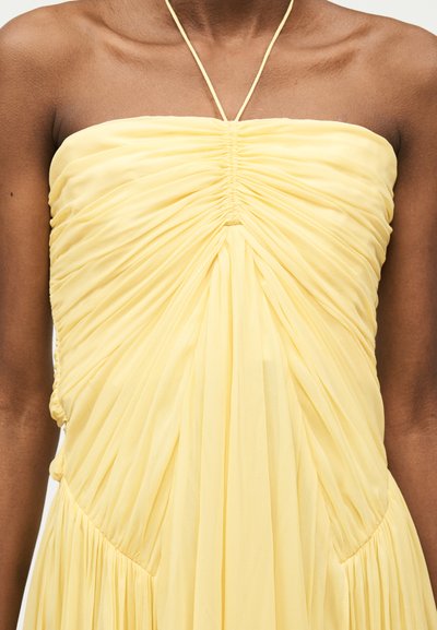 Žluté šaty bez ramínek s nařaseným živůtkem a halter výstřihem, zobrazené na horní části těla a pažích osoby na jednobarevném pozadí.