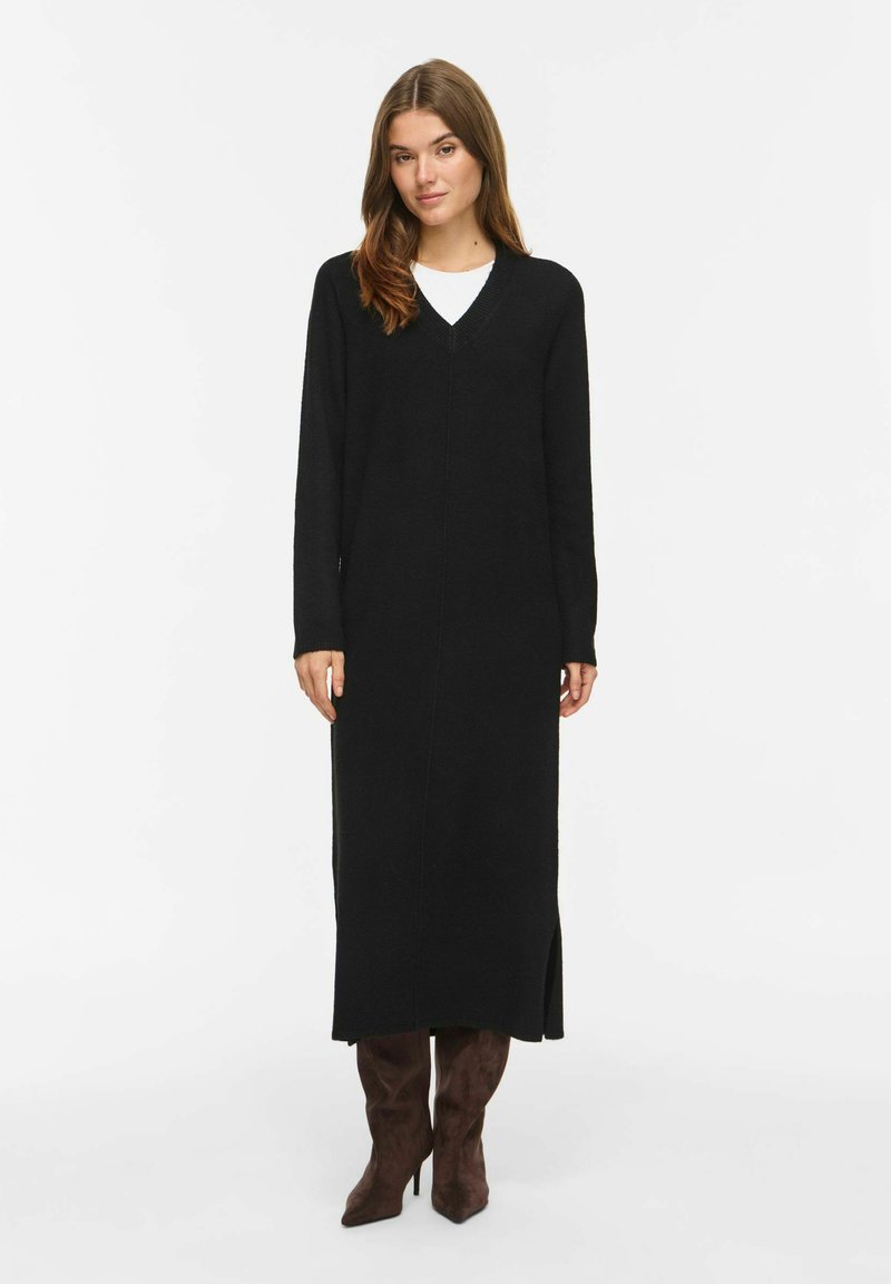 Femme aux longs cheveux bruns portant une longue robe noire à col en V et des bottes à talons marron foncé, debout devant un fond blanc uni.