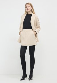 Femme portant un blazer et une jupe en velours côtelé beige avec un top côtelé noir, des collants noirs et des talons aiguille noirs, se tenant avec les mains dans les poches.