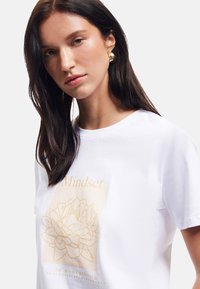 Camiseta de algodón blanca con gráfico floral dorado y texto. Mangas cortas, cuello redondo, corte relajado, textura suave, diseño moderno.