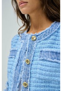 Ljusblå denimjacka med en strukturerad väv, fransade kanter, två framfickor och knäppningar med guldfärgad knapp.