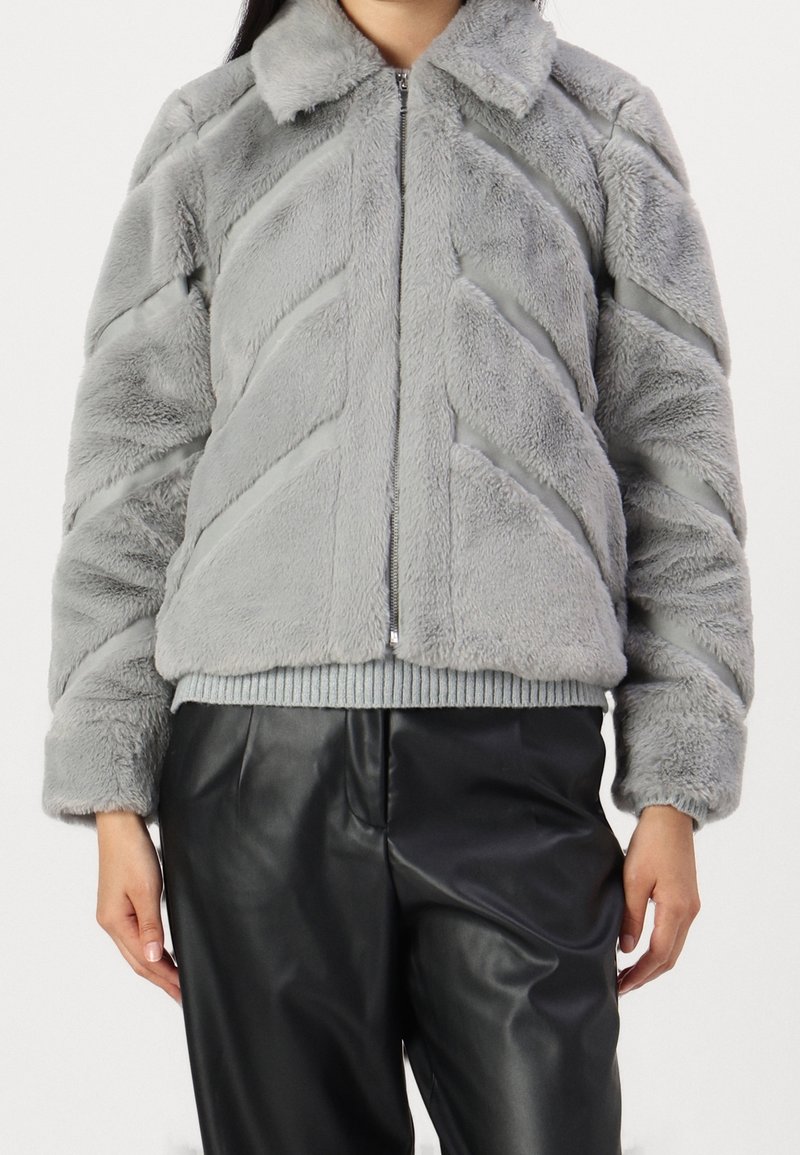 Giacca bomber in finta pelliccia grigia con motivo a chevron, chiusura con zip e colletto; polsini e orlo a coste; abbinata a pantaloni in finta pelle nera.