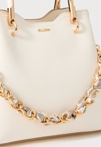 Bolso de mano de imitación cuero en color crema con herrajes en tono dorado y una correa de cadena trenzada que presenta acentos de tela estampada. Diseño con borde superior ondulado.