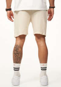 Beige strukturierte Shorts mit einem lockeren Schnitt, weiße Sneakers mit schwarzen Streifen und knöchellange Socken. Besitzt ein detailliertes Tattoo am Bein.