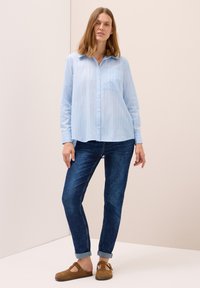 Camicia a righe blu chiaro con colletto e taschino frontale, abbinata a jeans blu scuro e scarpe con slip-on marroni.