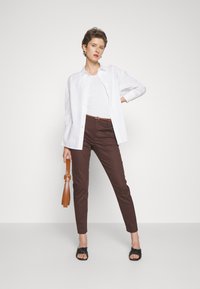 Chemise blanche à manches longues par-dessus un débardeur blanc, pantalon slim marron, sandales noires, et tenant un sac à main marron. Une tenue simple avec des lignes épurées.