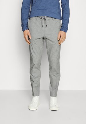 Mand iført lysegrå bukser med snøre, blå langærmet skjorte og hvide sneakers, stående mod en ensfarvet baggrund.