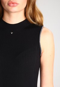 Svart ribbad ärmlös topp med hög krage och silverdetaljer längs axlarna, med en liten metallisk logotyp vid halsringningen.