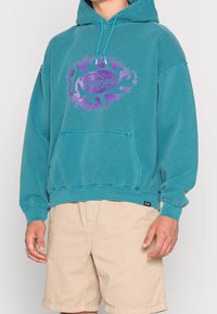 Tyrkysová mikina s kapucí z fleecu, s velkým fialovým grafickým motivem uprostřed, klokáním kapsou a nastavitelnou stahovací šňůrkou na kapuci.