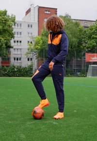 Donkerblauwe trainingspak met oranje accenten, gestructureerde stof, ritszakken, gedragen met feloranje schoenen, staand op groen kunstgras.