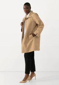 Cappotto beige con una vestibilità comoda, grandi bottoni, tasche laterali e un design pulito, abbinato a pantaloni neri e tacchi alti beige.