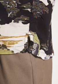 Bluzka z krótkim rękawem z nadrukiem kwiatowym w odcieniach czerni, zieleni i beżu. Dół ma boczne rozcięcia oraz widoczne detale szwów.