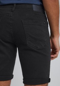 Black denim shorts featuring a rolled hem, subtle stitching, and two back pockets with a chevron design. Textured fabric.