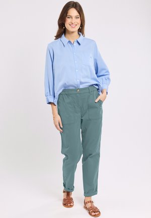 Smiling woman with brown hair wearing a light blue button-up shirt, teal pants with pockets, and brown woven sandals standing on white background.