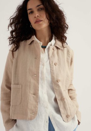 Naisel lokkis tumedate juustega, kandes beežikat nööpidega jopet valge särgi, siniste teksade ja türkiissinise kaelakeega ühevärvilise tausta ees.