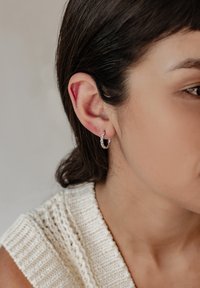 Orecchino a cerchio in argento con design intrecciato, caratterizzato da una finitura lucida. Inquadratura ravvicinata dell'orecchio con capelli corti e scuri e un top lavorato a maglia.