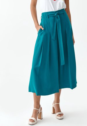 Vrouw draagt teal brede culottes met een tailleband, gecombineerd met witte sandalen met hoge hakken, staat met één hand in de zak.