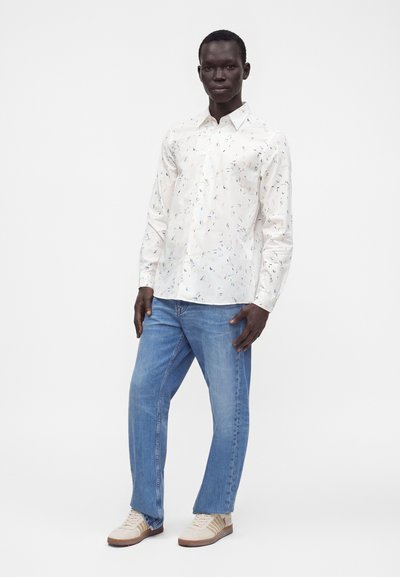 Homme debout portant une chemise blanche à manches longues avec de petites taches colorées, un jean bleu et des baskets beige sur un fond blanc uni.
