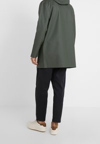 Osoba ubrana w zieloną wodoodporną kurtkę, czarne spodenki do kostek i białe tenisówki z brązowymi akcentami, stojąca na tle białego tła.