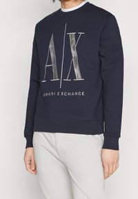 Felpa blu navy con maniche lunghe, polsini e bordo a coste. Presenta un logo bianco e argentato metallizzato "A/X" e la scritta "ARMANI EXCHANGE" sul davanti.