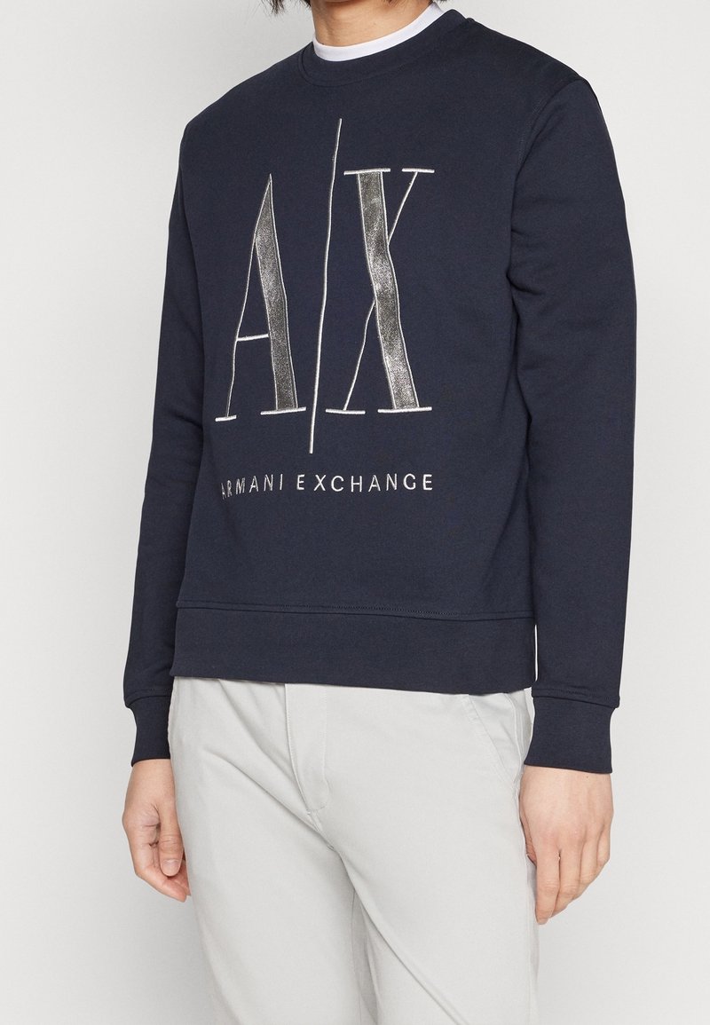 Felpa blu navy con maniche lunghe, polsini e bordo a coste. Presenta un logo bianco e argentato metallizzato "A/X" e la scritta "ARMANI EXCHANGE" sul davanti.
