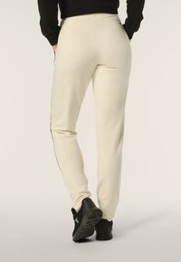 Pantalones de chándal beige con un ajuste slim, que cuentan con bolsillos laterales y una raya negra a lo largo de las costuras exteriores. Combinados con zapatillas deportivas negras.