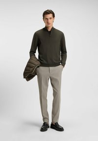 Khaki ritsvest met lange mouwen, gecombineerd met lichtgrijze broeken en zwarte instappers. Een bruine pufferjack is over één arm gegooid.