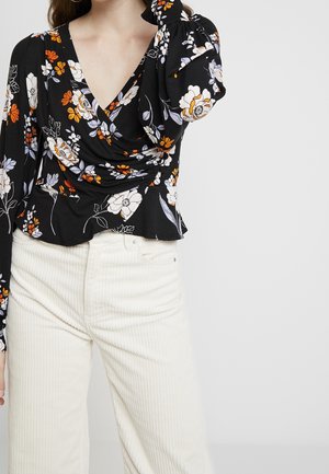 Frau trägt eine schwarze, langärmelige Blumenbluse mit Puffärmeln und weiße, hoch taillierte Cordhosen vor einfarbigem Hintergrund.