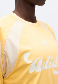 Άτομο που φοράει αθλητική μπλούζα Adidas σε κίτρινο και μπεζ χρώμα με ορατό λογότυπο και μεγάλο καλλιγραφικό κείμενο στο στήθος, εμφανιζόμενο από τον λαιμό έως τη μέση του κορμού.