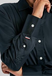 Svart skjorta med knappar och långa ärmar, som har vita knappar, en jämn textur och en liten röd, vit och blå detalj p å ärmsluten.