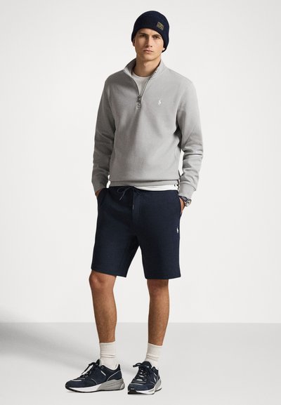 Άνδρας που φοράει ναυτικό σκουφάκι, ανοιχτό γκρι πουλόβερ με φερμουάρ στο λαιμό, ναυτικά σορτς, λευκές κάλτσες και ναυτικά αθλητικά παπούτσια, στέκεται με τα χέρια στις τσέπες.