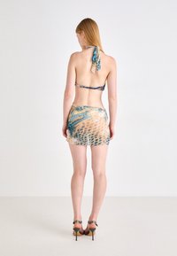 Bikini top s halter dekolteom, uzorom u nijansama plave i bež, uparen s odgovarajućom, prozirnom mini suknjom. Sandale s visokom petom.