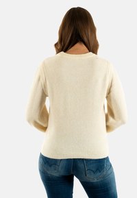 Suéter de punto color crema con cuello redondo y mangas largas, que presenta un acabado texturizado y puños y dobladillo acanalados. Llevado con jeans azules.