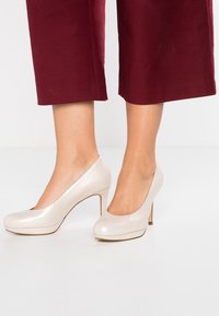 Donna che indossa eleganti décolleté off-white lucide abbinati a pantaloni cropped rossi scuri, in piedi su un pavimento chiaro.