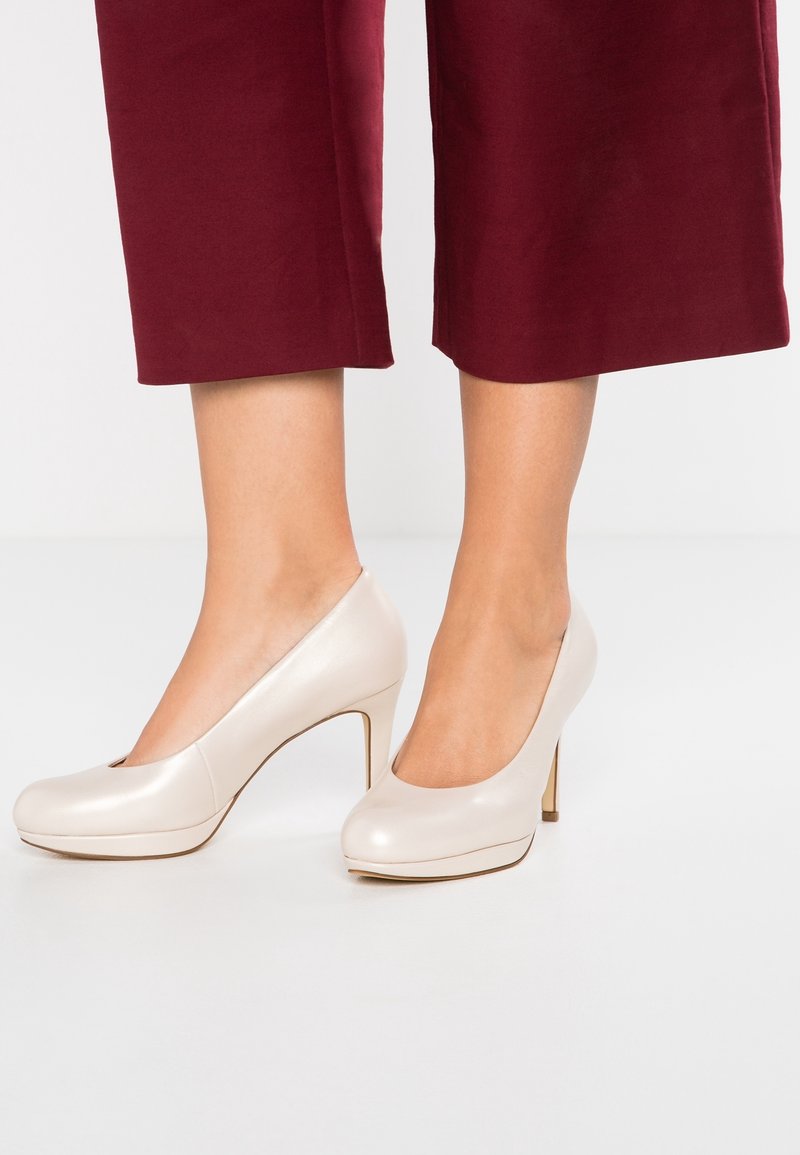 Donna che indossa eleganti décolleté off-white lucide abbinati a pantaloni cropped rossi scuri, in piedi su un pavimento chiaro.