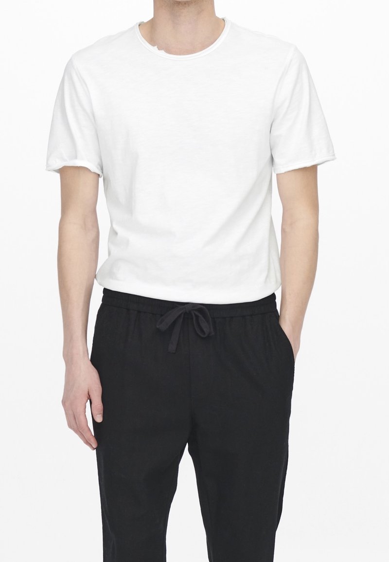 Homme portant un t-shirt blanc uni à manches courtes, rentré dans un pantalon noir à cordon, les mains détendues le long du corps.