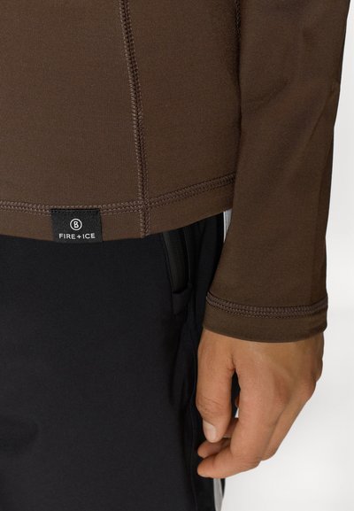 Κοντινό πλάνο ενός χεριού δίπλα σε ένα καφέ μακρυμάνικο πουκάμισο με ετικέτα "Φωτιά + Πάγος" πάνω από μαύρα παντελόνια.