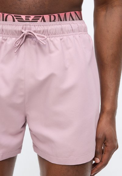 Άνδρας που φοράει ανοιχτό ροζ σορτς με κορδόνι, με ορατή ροζ ζώνη μέσης που φέρει το λογότυπο "Armani".
