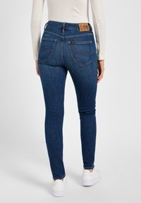 Jean skinny en denim bleu avec une taille haute, présentant une étiquette en cuir marron à l'arrière et deux poches, associés à des baskets blanches.