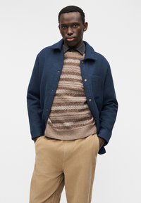 Μπλε ναυτικό μάλλινο σακάκι με γιακά, εμπρόσθια κουμπιά και τσέπη στο στήθος πάνω από πλεκτό καφέ και μπεζ ριγέ πουλόβερ. Μπεζ παντελόνι από κοτλέ.