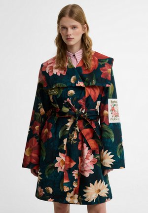 Γυναίκα που φοράει σκούρο φλοράλ παλτό με ζώνη, με μεγάλα κόκκινα, ροζ και λευκά λουλούδια και ένα μπαλώμα στο μανίκι που γράφει "Barbour Farm Rio."