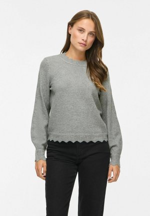 Femme aux longs cheveux bruns portant un pull gris à bords festonnés et un pantalon noir, debout sur un fond blanc uni.